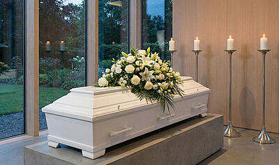 Weißer Sarg mit Blumenschmuck. Kerzen im Hintergrund. Links Blick durch Fenster auf einen Garten