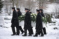 6 Männer tragen einen weißen Sarg mit Tannengesteck. Es liegt Schnee.