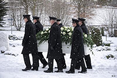 6 Männer tragen einen weißen Sarg mit Tannengesteck. Es liegt Schnee.